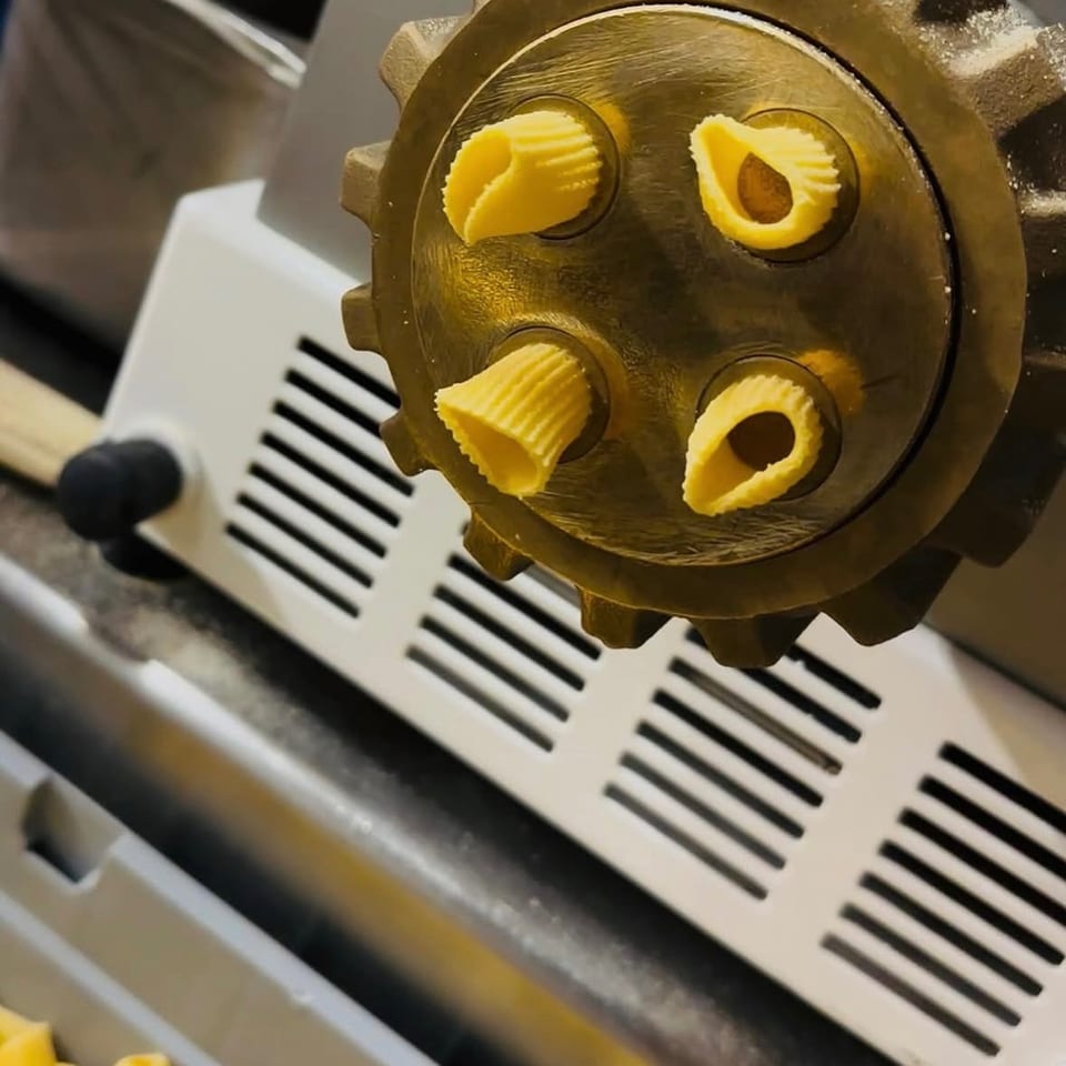 Freshly extruded pasta, shaped like small shells, emerging from a pasta machine.