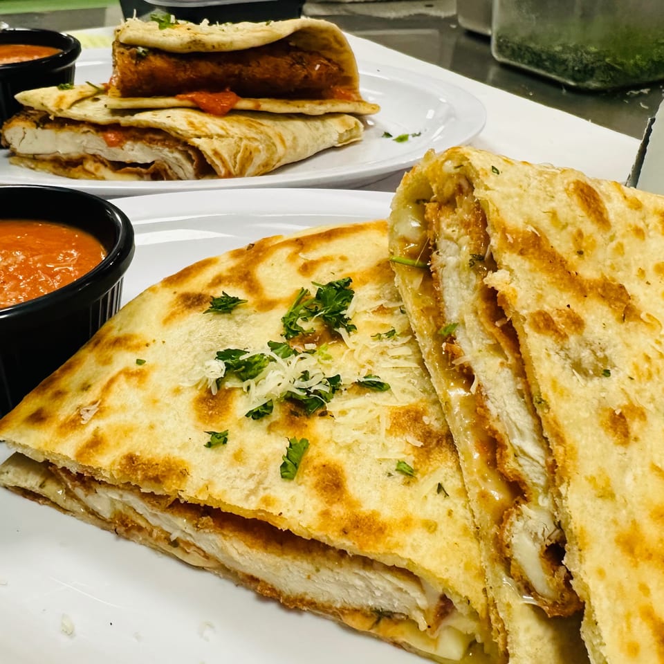 Two plates of chicken quesadillas with melted cheese, garnished with parsley, and served with red dipping sauce.