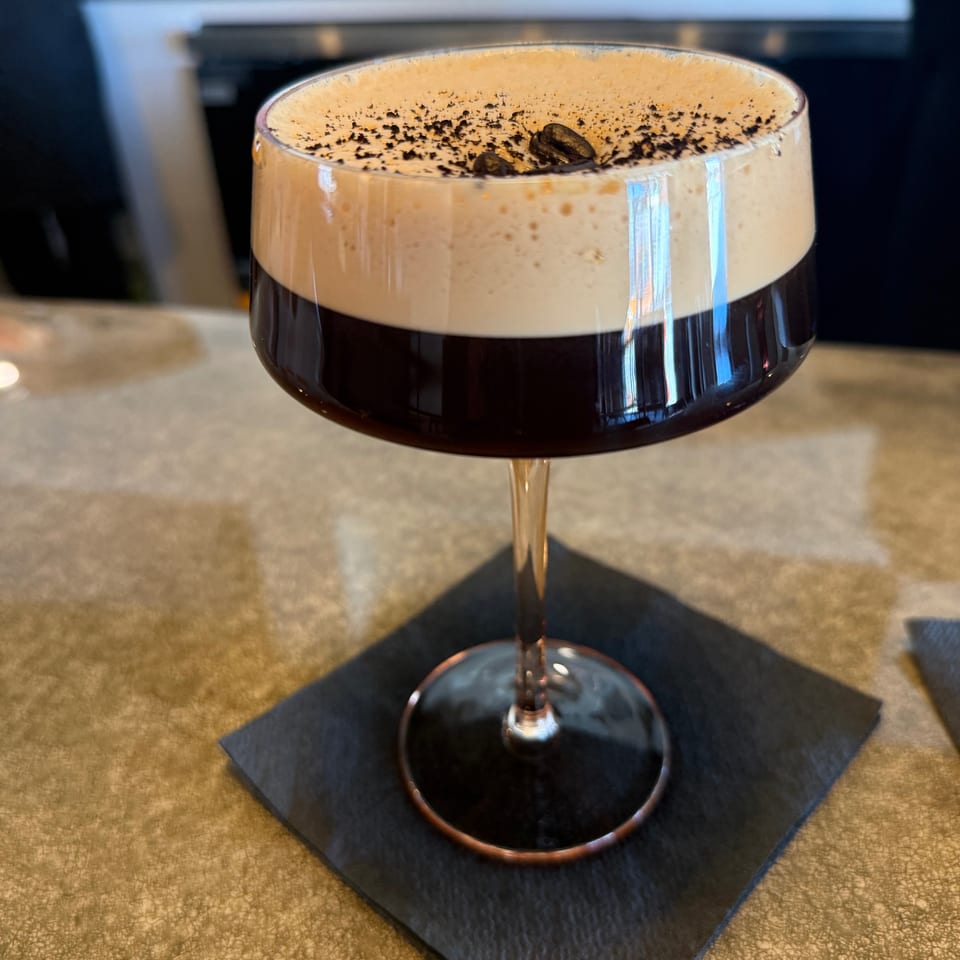 Espresso martini in a coupe glass with foamy top, coffee beans, and cocoa powder on a black napkin.